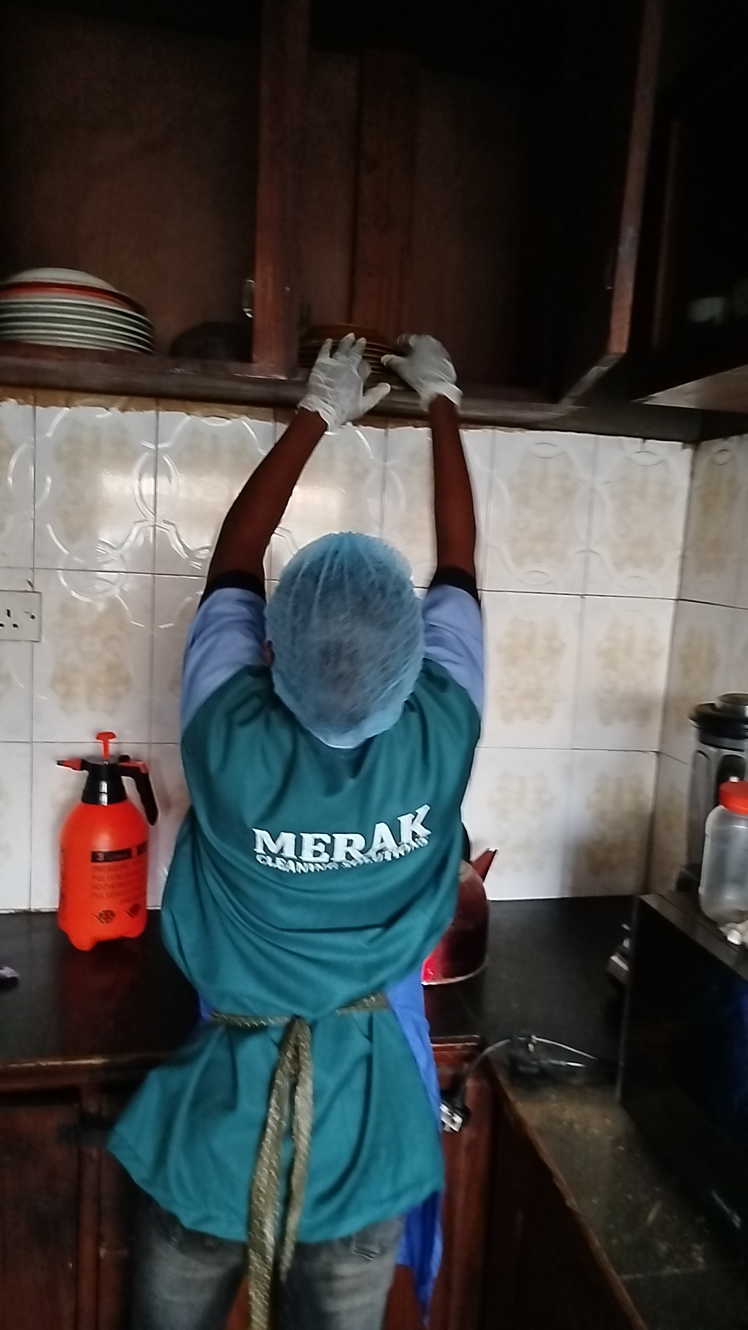 Merak Cleaning Solutions residential cleaner in branded uniform deep-cleaning kitchen cabinets and shelves in a Nigerian home